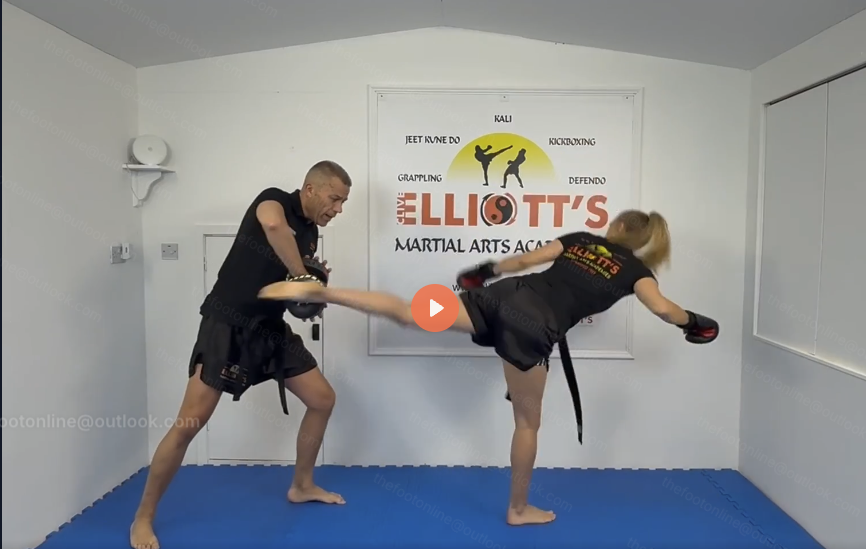 Clive Elliott holding a pad while Kaz throws a high roundhouse kick in the academy dojo.